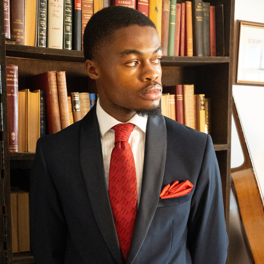 A man wearing a navy blue suit jacket, white dress shirt, and a bright red patterned tie with a matching red pocket square, standing in front of a bookshelf.